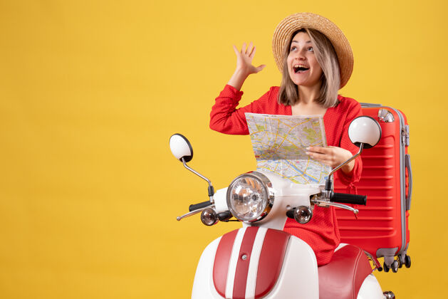 坐着骑着轻便摩托车的漂亮女孩拿着红色的手提箱 举着地图向上看时尚红色地图