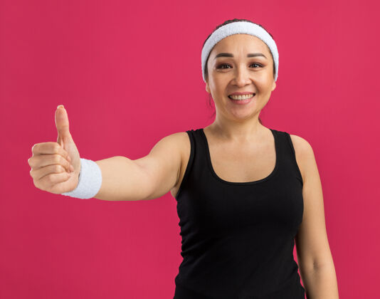 微笑戴着头巾的年轻健身女士站在粉红色的墙上愉快地笑着竖起大拇指欢呼拇指表演