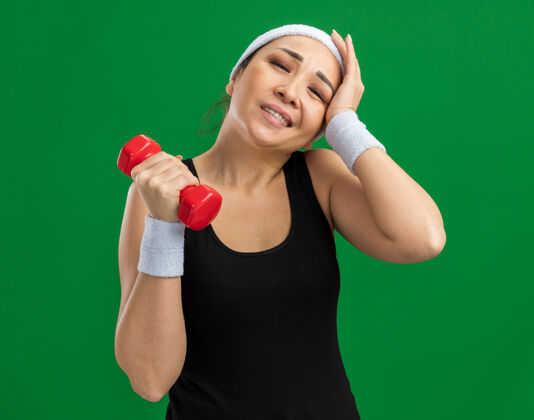站立戴着哑铃的头带的年轻健身女士站在绿色的墙上做着看起来很累很累的运动健身头带运动
