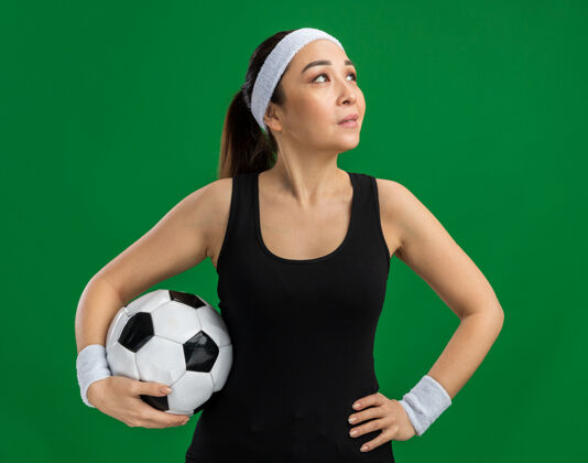 年轻戴着头巾 手持足球 面带微笑的年轻健身女士站在绿色的墙上微笑脸头带