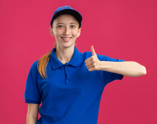 女孩身穿蓝色制服 头戴鸭舌帽的年轻送货女孩站在粉红色的墙上 自信地微笑着竖起大拇指交货立场拇指