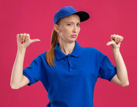 站着穿着蓝色制服 戴着帽子的年轻送货女孩站在粉红色的墙上 带着怀疑的表情指着自己帽子交货年轻