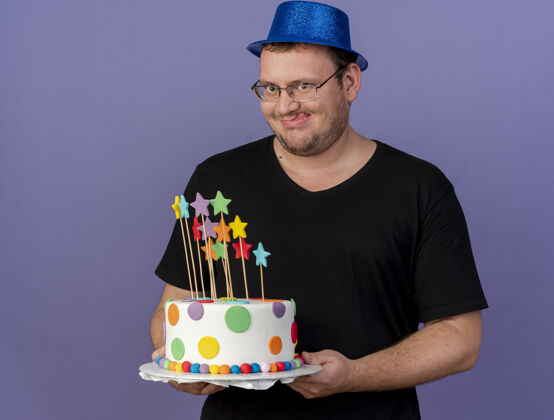 蛋糕兴奋的成年斯拉夫人戴着眼镜 戴着蓝色派对帽 伸出舌头 手里拿着生日蛋糕卡住生日聚会