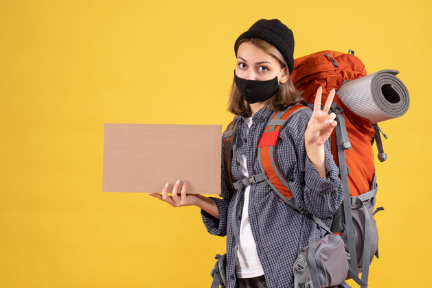 人带着黑色面具和背包的旅行女孩拿着纸板制作胜利标志女孩旅行者帽子纸板