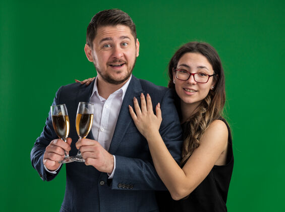 眼镜年轻漂亮的夫妇幸福的男人 带着香槟和微笑的女人 拥抱着幸福的爱情 一起庆祝情人节站在绿色的墙上香槟微笑一天