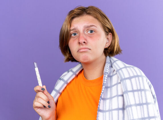 感觉不健康的年轻女子裹着温暖的毯子 感觉不舒服 患流感 发烧 用温度计测量体温 站在紫色的墙上 看起来很担心痛苦女人担心