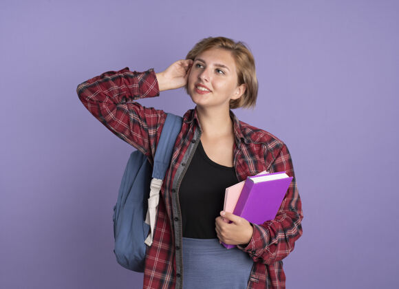 紫色高兴的年轻斯拉夫学生女孩戴着背包 手放在头上 拿着书和笔记本看着身边年轻背包空间