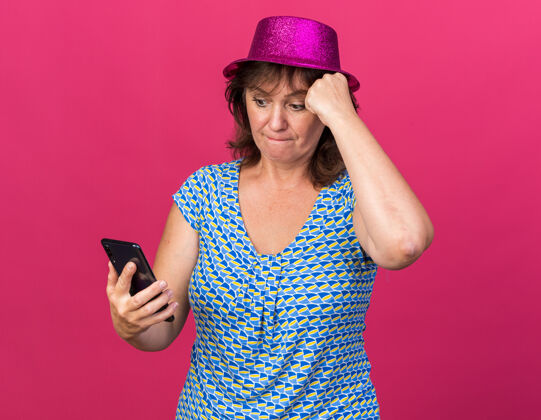 女人戴着派对帽的中年妇女看着她的智能手机屏幕困惑地站在粉色的墙上庆祝生日派对困惑中间手机