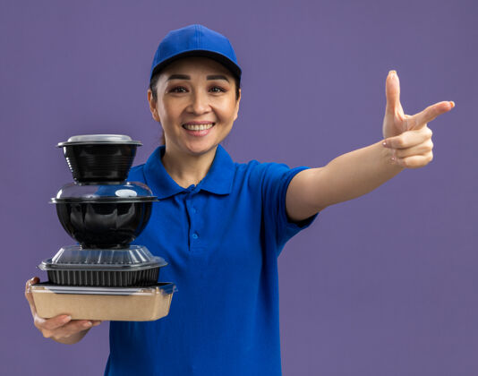 拿着身穿蓝色制服 头戴鸭舌帽的年轻送货员手里拿着一叠食品包 愉快地微笑着 食指站在紫色的墙上盒子送货帽子