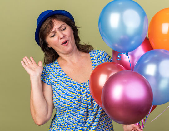 生日快乐的中年妇女戴着派对帽 手里拿着一堆五颜六色的气球 站在绿色的墙上欢庆生日派对立场乐趣五颜六色