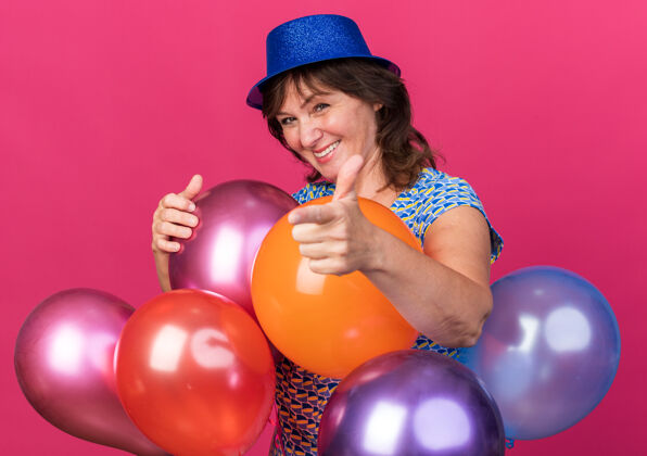 指向戴着派对帽的中年妇女手持五颜六色的气球 开心地微笑着 用食指指着紫色的墙上 庆祝生日派对目录立场生日