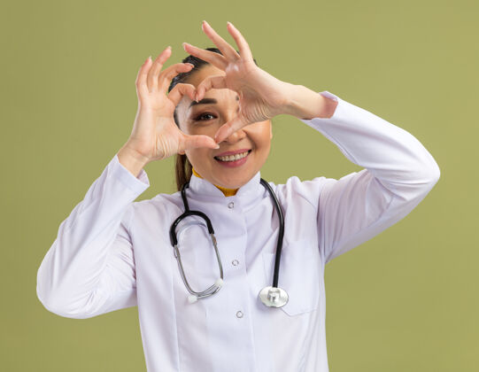 女人年轻的女医生 穿着白药衣 脖子上戴着听诊器 站在绿色的墙上做着用手指看着的心脏手势做微笑通过
