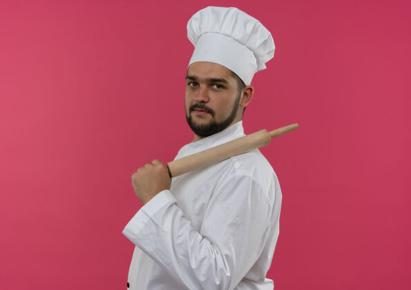 肩膀自信的年轻男厨师身着厨师制服站在侧视图上 将擀面杖放在肩膀上 隔离在粉色墙壁上 留有复制空间观点别针信心