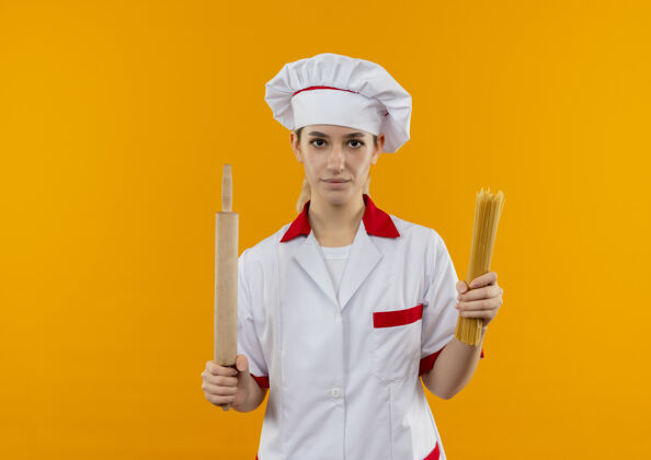 烹饪自信的年轻漂亮厨师穿着厨师制服 拿着意大利面和擀面杖 隔离在橙色的墙上 留有复制空间制服厨师意大利面