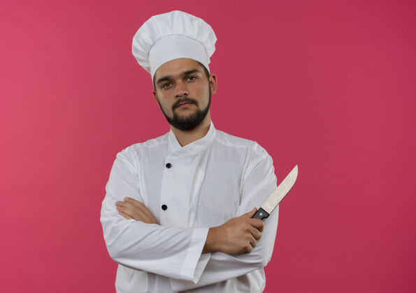 姿势自信的年轻男性厨师身着厨师制服 站在粉红色的墙上 保持封闭的姿势 手持隔离的刀年轻站立封闭