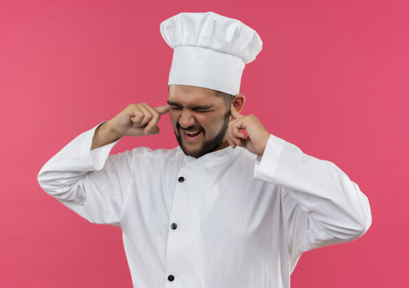 男性恼怒的年轻男厨师穿着厨师制服把手指放在耳朵里闭着眼睛孤立在粉红色的墙上烹饪恼火闭嘴