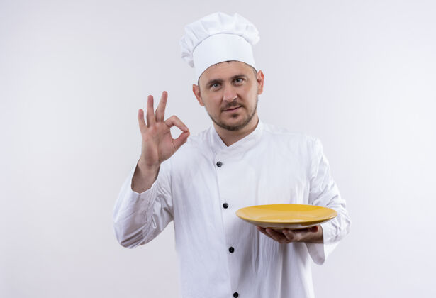 烹饪自信的年轻帅哥厨师穿着厨师制服拿着盘子 在孤零零的白墙上做着ok的手势制服厨师帅气