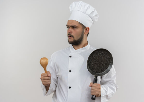 年轻印象深刻的年轻男厨师在厨师制服举行汤匙和煎锅隔离在白色的墙壁与复制空间勺子烹饪薯条