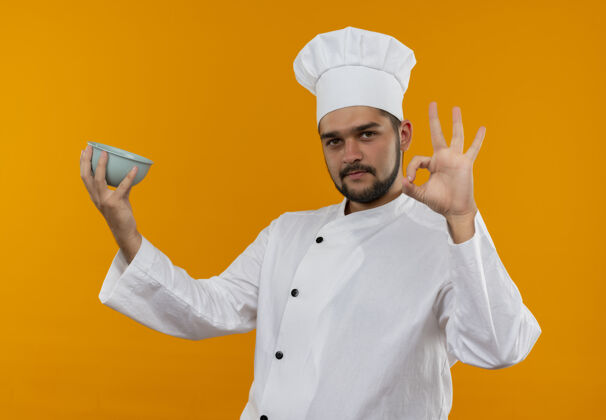 好自信的年轻男厨师身着厨师制服 拿着碗 在橙色的墙上贴着“一切正常”的牌子烹饪碗男性