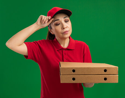 送货身穿红色制服 头戴鸭舌帽 拿着披萨盒的年轻送货员站在绿色的墙上 神情悲伤地低头看着悲伤站着帽子