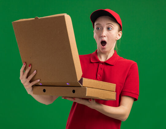 拿着身穿红色制服 头戴鸭舌帽的年轻送货女手里拿着披萨盒 站在绿色的墙壁上 打开其中一个披萨盒 我既惊讶又惊讶送货打开惊喜