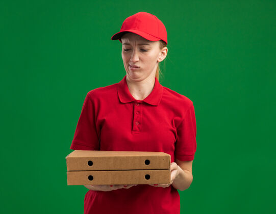 年轻年轻的送货员身穿红色制服 头戴鸭舌帽 手里拿着披萨盒 站在绿色的墙上 困惑而不高兴地看着它们不高兴帽子盒子