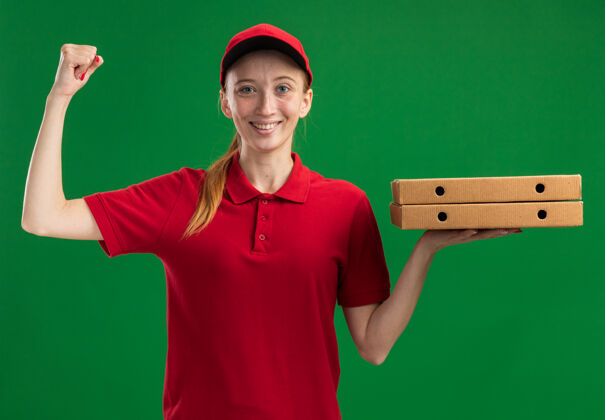 拳头身穿红色制服 头戴鸭舌帽 手拿披萨盒的年轻送货女孩微笑着自信地举起拳头站在绿色的墙上帽子信心抱着