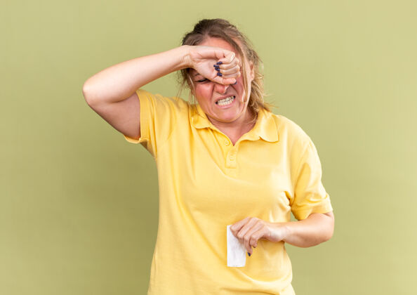 触摸不健康的穿黄衬衫的女人摸额头感觉很难受 因为鼻子堵了 感冒站在绿色的墙上 头痛得厉害鼻子可怕女人