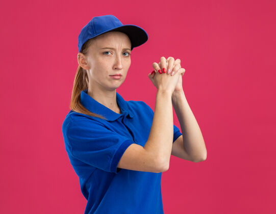 女孩身穿蓝色制服 头戴鸭舌帽的年轻送货员 表情严肃自信 手牵手 站在粉色的墙上 做出团队合作的姿态年轻团队合作制服