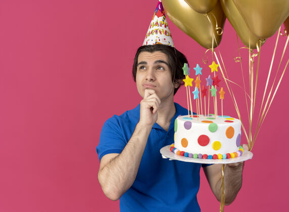 手困惑英俊的白种人戴着生日帽 手放在下巴上 手里拿着氦气球和生日蛋糕迷糊空格拿着