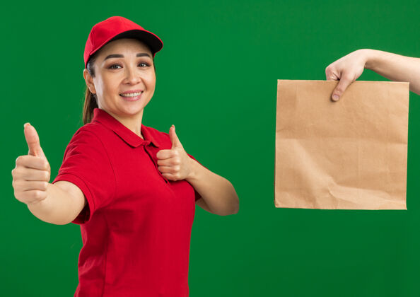 帽子年轻的送货员身穿红色制服 头戴鸭舌帽 微笑着竖起大拇指 站在绿色的墙边收包裹站立送货同时