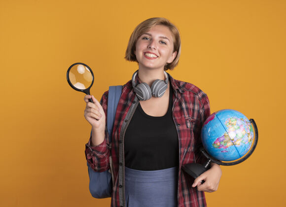 拿着微笑的年轻斯拉夫女学生戴着耳机背着背包拿着放大镜和地球仪微笑复制耳机