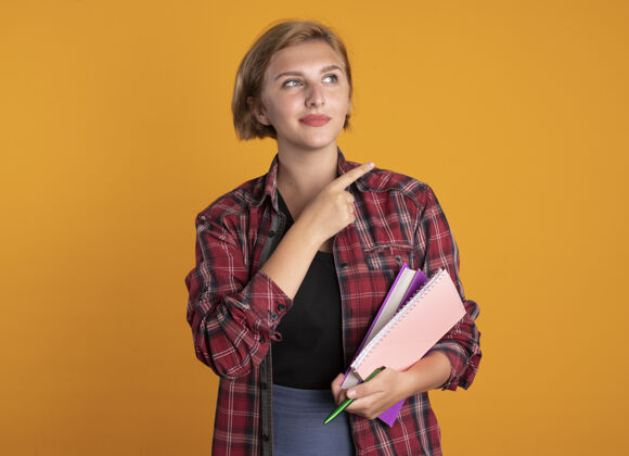 复制高兴的年轻斯拉夫学生女孩拿着笔本和笔记本看着并指着身边钢笔空间橙色