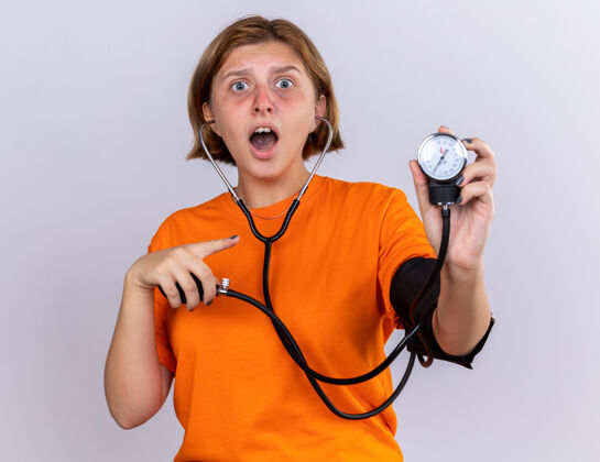 血压计不健康的年轻女子穿着橙色t恤 感觉不舒服 用血压计测量血压 站在白色的墙上 看起来很担心站立不健康不适