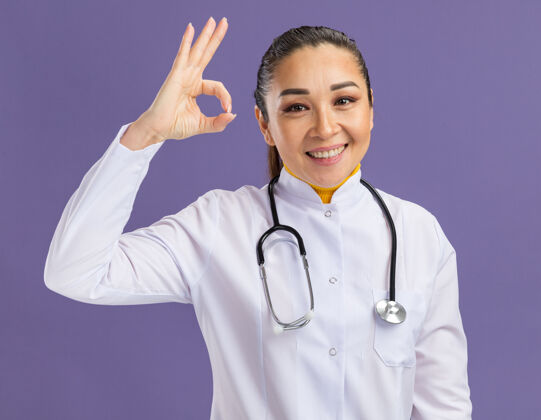 女人身穿白色药衣 脖子上戴着听诊器 脸上带着微笑的年轻女医生站在紫色的墙上做着“ok”的手势年轻脸听诊器