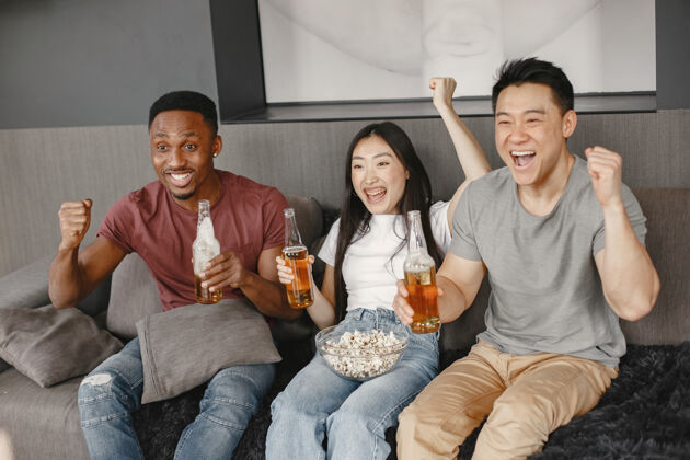 积极非洲男孩和亚洲夫妇用啤酒碰杯朋友们看足球赛 吃爆米花人们为足球队加油时间娱乐房子