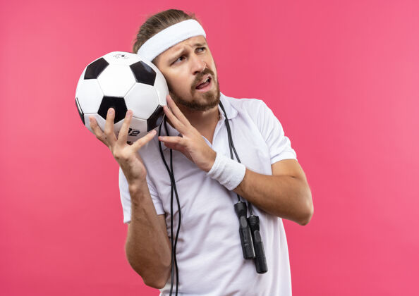 帅气困惑的年轻英俊的运动型男子戴着头带和手环拿着足球看一边跳绳围着脖子隔离在粉红色的墙上球头带侧身