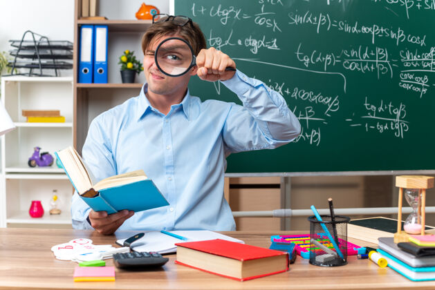 坐着年轻的男老师戴着眼镜透过放大镜看着教室里的黑板前 坐在课桌旁的书本和笔记眼镜老师玻璃