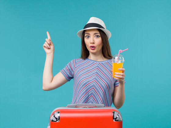 航行前视图：度假中的女人 在浅蓝色的背景下提着她的红色袋子 手里拿着果汁 旅途中 旅途中 度假中的女人 旅途中休闲人旅行