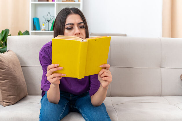 举行穿着休闲服的年轻女孩抱着书严肃地坐在客厅的沙发上看书阅读年轻坐