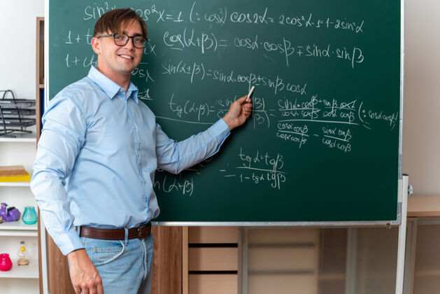 课年轻的男老师戴着眼镜拿着粉笔讲课 微笑着自信地站在教室里数学公式的黑板旁数学年轻人粉笔