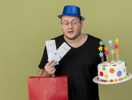 机票令人印象深刻的成年斯拉夫人戴着眼镜戴着蓝色派对帽拿着纸购物袋生日蛋糕和机票生日航空橄榄色