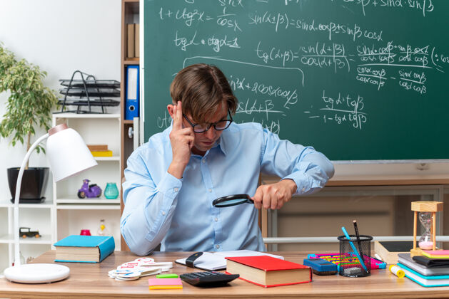 年轻年轻的男老师戴着眼镜通过放大镜看笔记备课坐在课桌旁 教室的黑板前放着书和笔记男人数学学校