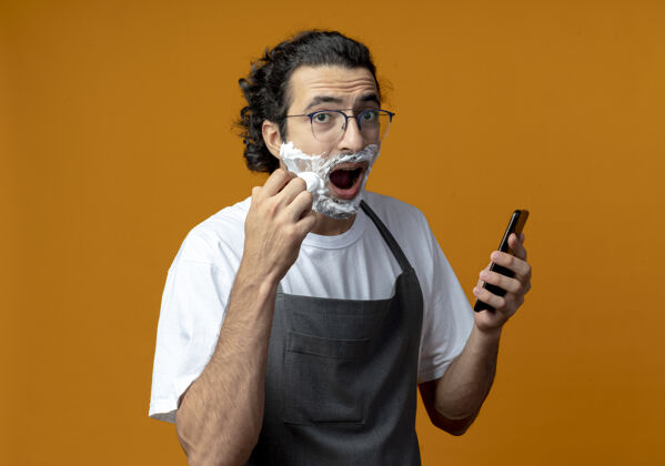 理发师印象深刻的年轻白人男性理发师戴着眼镜和波浪发带制服拿着手机 把剃须膏抹在脸上橙色男性手机