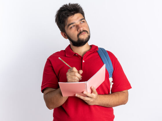 表情年轻的留着胡子的学生 穿着红色马球衫 背着书包 在笔记本上写字 抬起头沉思着站着向上学生胡子