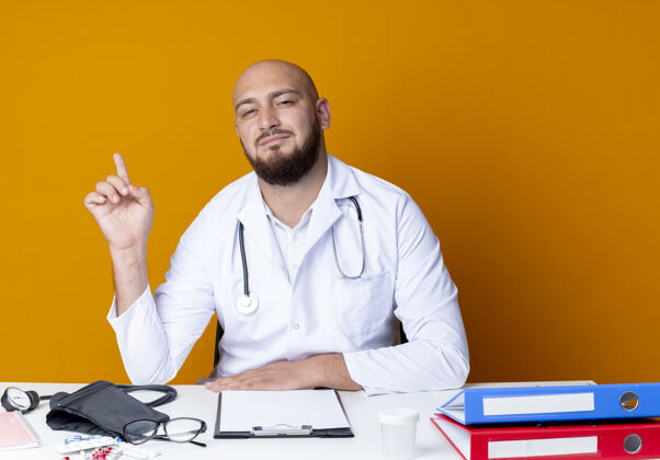 男性高兴的年轻秃头男医生穿着医用长袍和听诊器坐在办公桌旁工作听诊器年轻
