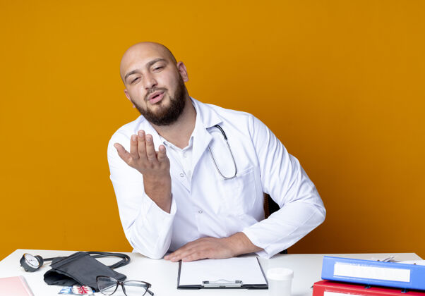 坐着年轻的秃头男医生穿着医用长袍和听诊器坐在办公桌旁工具接吻医疗