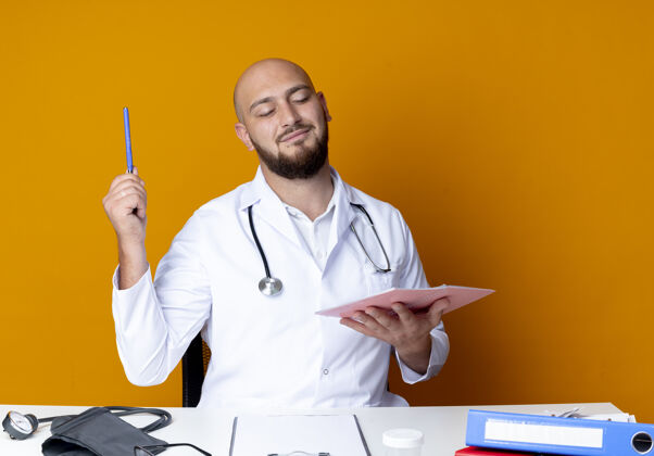 坐着高兴的年轻秃头男医生穿着医用长袍和听诊器坐在办公桌旁钢笔橙色请穿