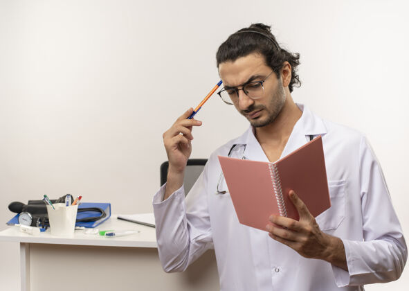 前面年轻的男医生戴着医用眼镜 穿着医用长袍 手持听诊器站在办公桌前听诊器桌子男性