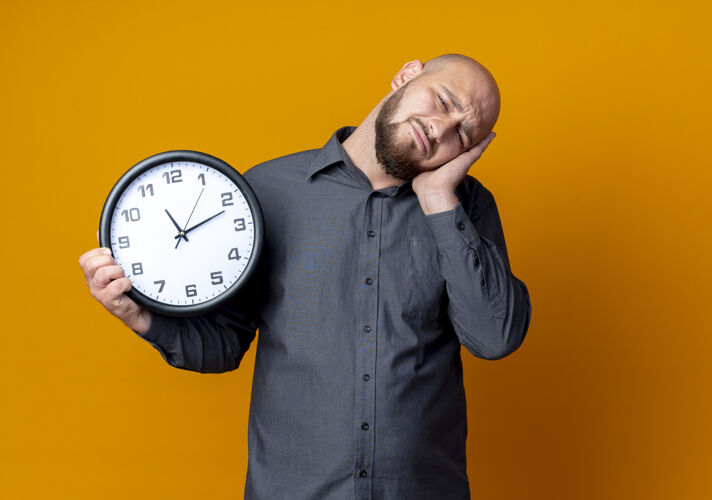 表情疲惫的年轻秃头呼叫中心男子拿着时钟 做着睡觉的姿势孤立在橙色的墙上疲倦握着秃头